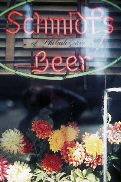 Exterior view of neon Schmidt's Beer sign in a diner window, along with an arrangement of flowers.