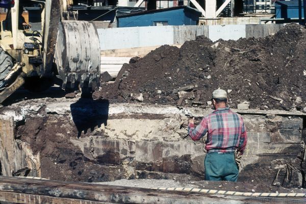 A male construction worker stands stands in a large hole directing another man who is using a backhoe.