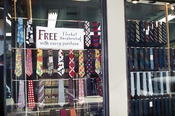 Exterior view of a shop's window display of various ties. A sign reads, "Free pocket handkerchief with every purchase."