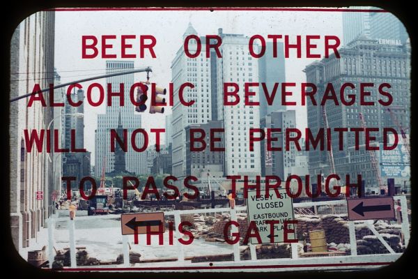A notification is on a window that reads, "Beer or other alcoholic beverages will not be permitted to pass through this gate." Beyond the gate, the World Trade Center construction site is visible. St. Paul's Chapel is in the far background.