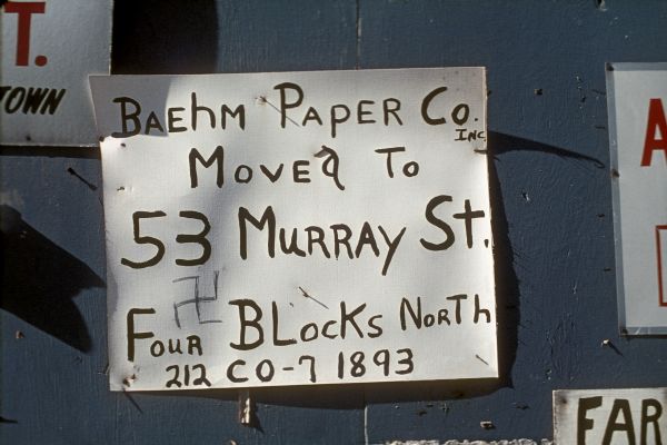 Close-up of a handmade sign reading: "Baehm Paper Co. moved to 53 Murray St. Four blocks north, 212 CO-7 1893." Someone has added a graffiti swastika on the sign.