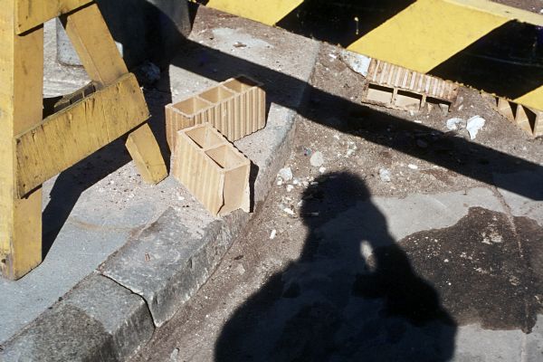 A shadow of the photographer, Richard Quinney, is visible on the street near the construction site. On the curb are cement blocks and sawhorses.