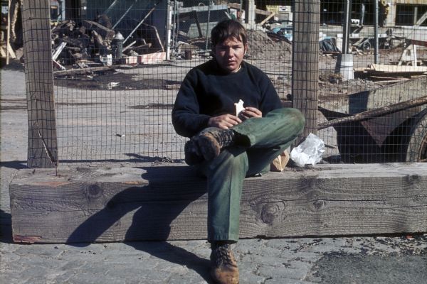 A construction worker sits on a wooden beam near a fence and eats his lunch. Behind him is the construction site with debris.