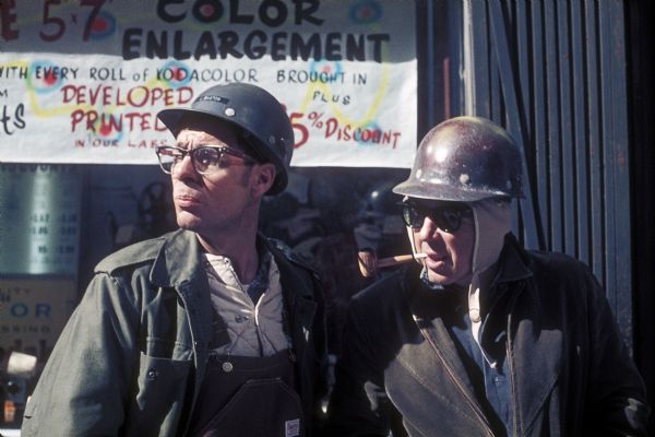 Two construction workers wearing hardhats stand outside a storefront while on a break.
