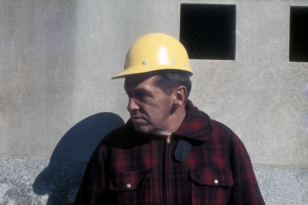 Close-up of a construction worker near a brick wall. He is dressed in a black and red flannel shirt and wears a yellow hard hat.