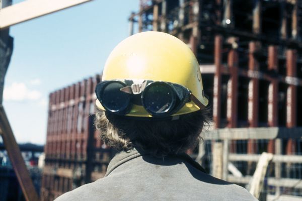 Rear view of a construction worker wearing safety goggles on the back of his yellow hard hat. In the background is the steel framework of one of the towers of The World Trade Center.