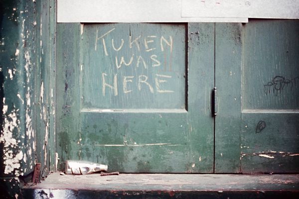 Graffiti on a green door reads, "Kuken was here." A Miller beer bottle is on the doorstep.