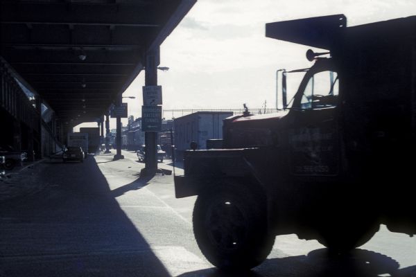 A man is driving a construction truck in the shadow cast by what appears to be an elevated highway. A sign for the "Brooklyn Battery Tunnel" is on one of the support columns of the elevated highway. In the background are trucks and buildings along a street.