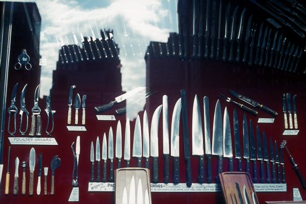 Knives and shears in a display case behind a show window, which is reflecting buildings and clouds in the sky.