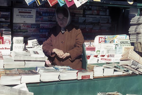 A woman stands behind the counter under an awning of a newspaper stand.