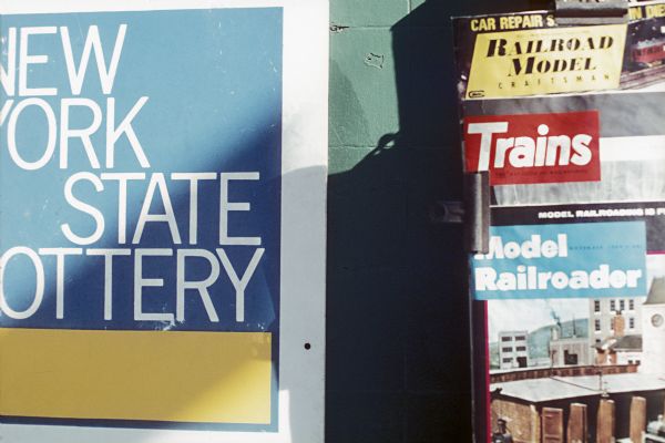 On the left is part of a "New York State Lottery" sign, and on the right, a number of magazines are displayed, probably part of a newspaper stand.