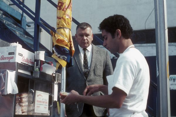 A man in a suit waits for a hot dog that a man is preparing. Metal steps painted blue rise behind the customer on the left.