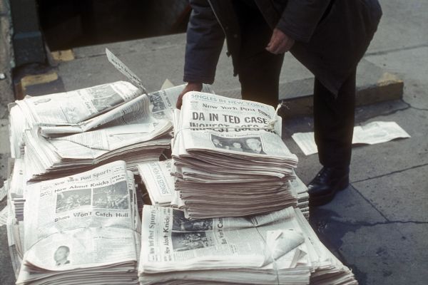 A man bends over stacks of sections of the <i>New York Post</i> piled on a sidewalk.