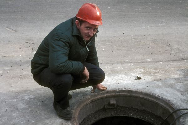 A construction worker wearing a red hard hat crouches next to an open manhole in the street.