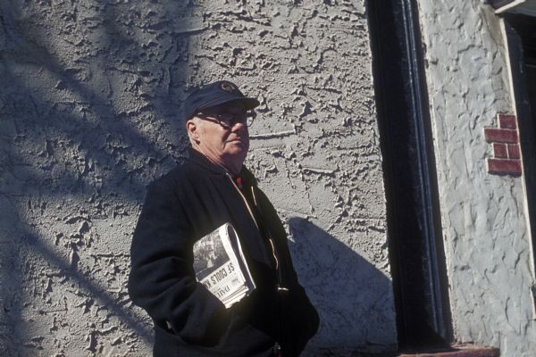 A man wearing a hat stands near a wall with his hands in his jacket pockets, holding a newspaper under his arm.