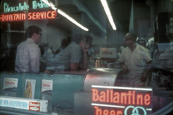View through window of a diner of several men engaged in conversation at the counter. A neon sign in the window says, in part: "Fountain Service."