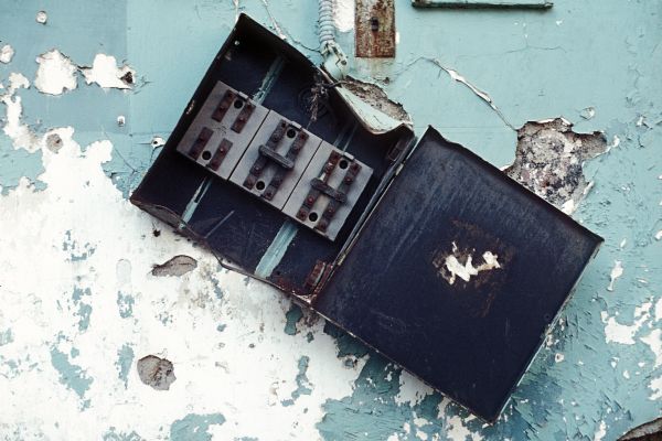 View of a broken electrical box hanging from the side of a building with peeling paint.