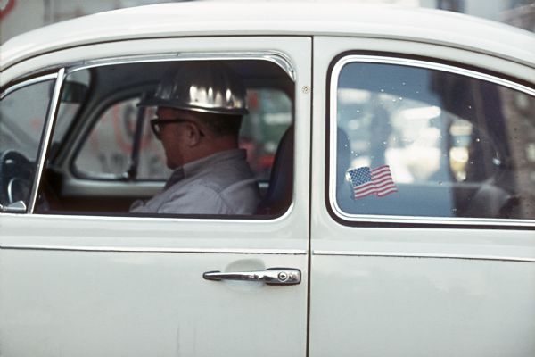 A construction worker wearing a silver hard hat sits in the driver's seat of a Volkswagen. An American flag sticker is visible on the backseat window.