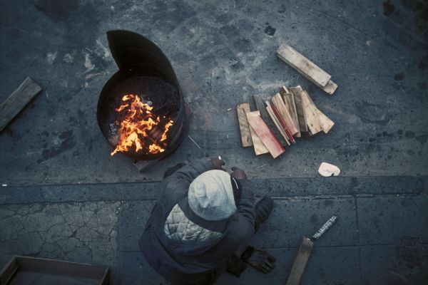 Overhead view of a homeless man sitting by a fire in a large barrel.