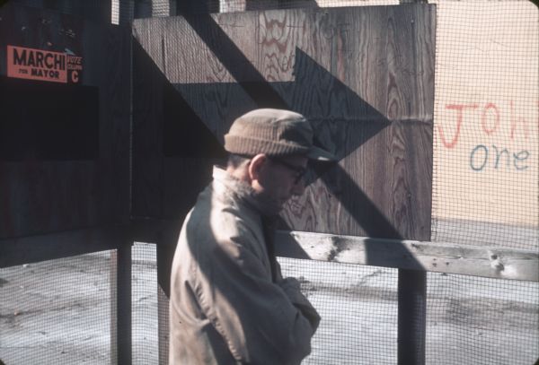 A man in the foreground walks past a wire fence with plywood at the construction site. A sign in the background reads, "Marchi for Mayor," and the portion of graffiti that is visible reads, "Joh(n) One."