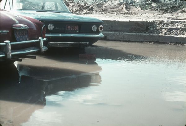 A large puddle in a parking lot where two automobiles, a Corvair and a Volkswagen, are parked.