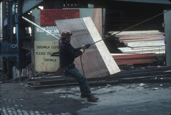 A construction worker leans back while pulling on a large rope. Construction equipment, supplies, and signs are in the background.