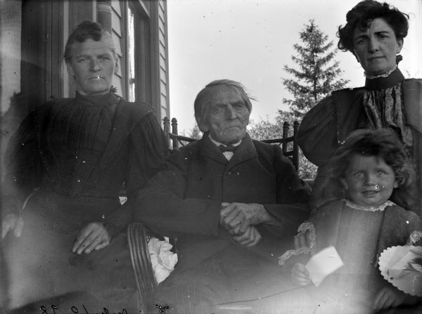 Syl sits outside on a porch with three other well-dressed family members of varying ages, each representing a generation in the family. Carl Schmidt is the elderly gentleman in the middle.