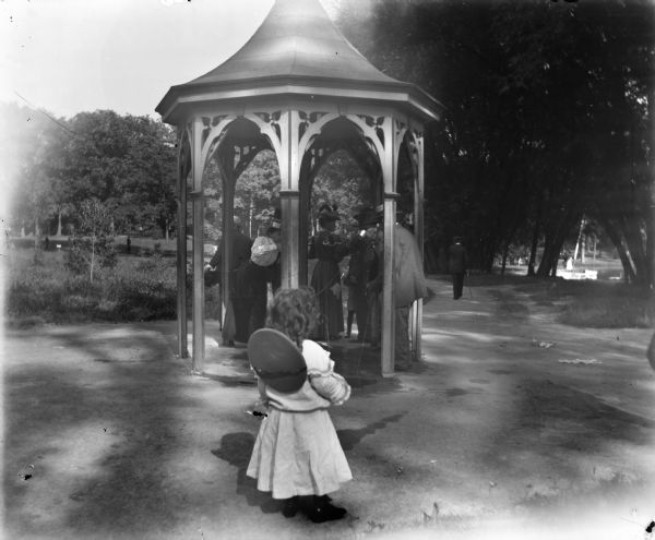 Syl stands in front of a pavilion with a well or water pump in a park. A group of adults dressed fashionably stand in the pavilion while drinking water from small cups. In the background are paths, trees, and a pond.