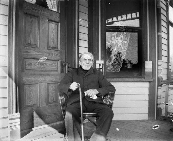 Carl Schmidt seated in a chair with a cane on a porch in front of a door and a window.