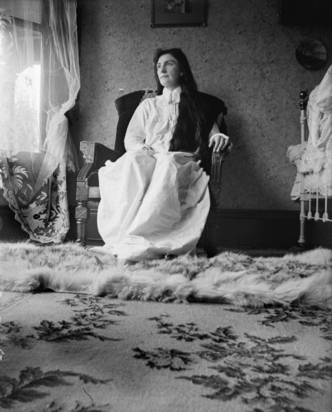 Low angle view of "Ma" sitting in a large chair near a window wearing a long white dress or nightgown in a bedroom with a fur rug over another rug. There are framed pictures on the wall and a bed on the right.