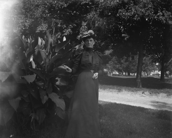 "Ma" posing near plants at Esou's. She is wearing a long dress and a fancy hat with feathers. A gravel road and other vegetation are in the background.