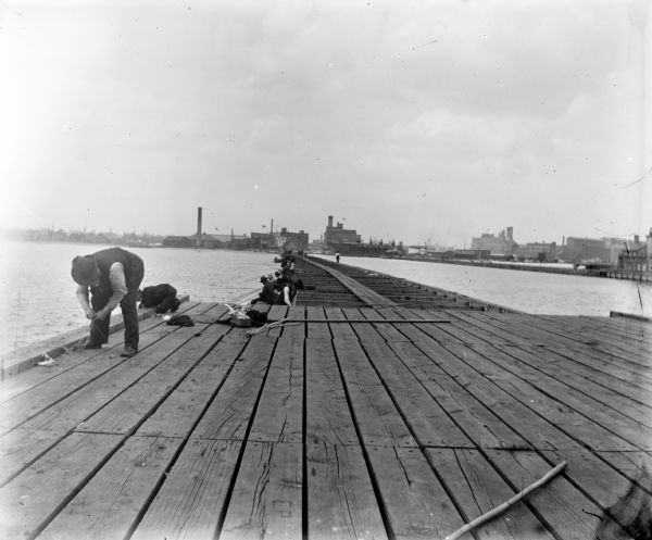 Fishermen fishing off of a long pier. Industrial buildings are across the water in the background.