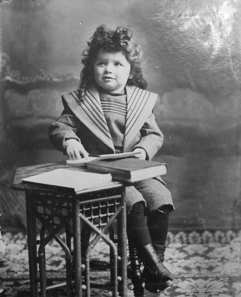 A portrait of Syl with long curly hair sitting next to a small table  with books on top of it.