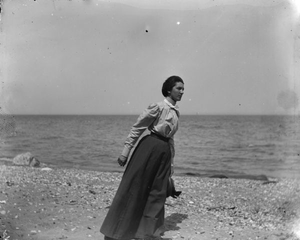 Aunt Helen leaning into the wind while holding a hat in her left hand and walking barefoot on the shore of a lake.