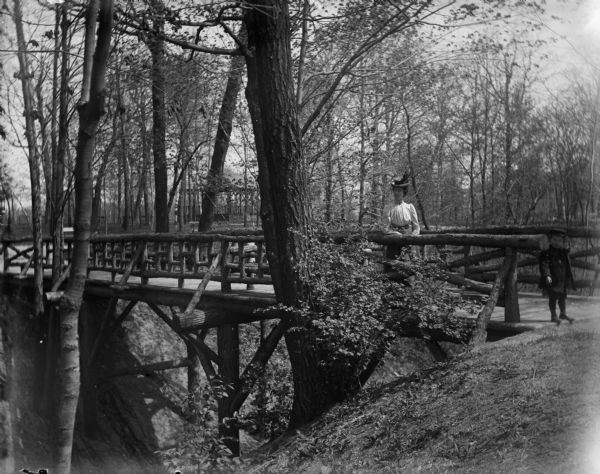 Aunt Helen and Syl stand on a rustic wooden bridge in Gully Lake Park. A pavilion is visible behind them through the trees.