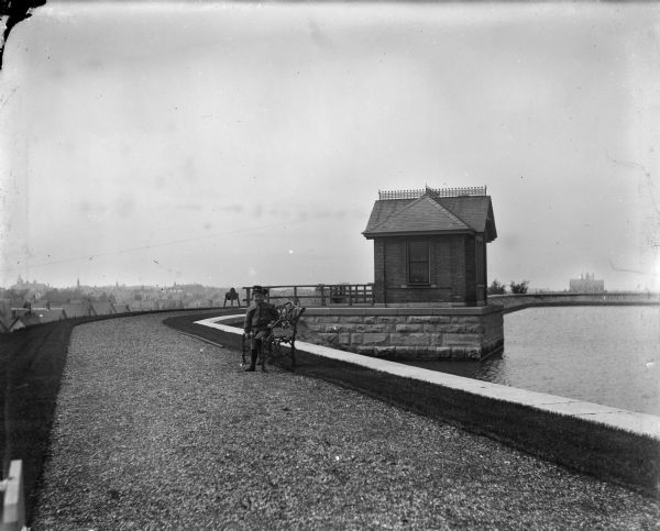 View down gravel path towards Syl seated on a bench along the edge of a reservoir on a hill. There is a brick building, perhaps a pumphouse, built over the water behind him. In the far distance are rooftops and a church building.