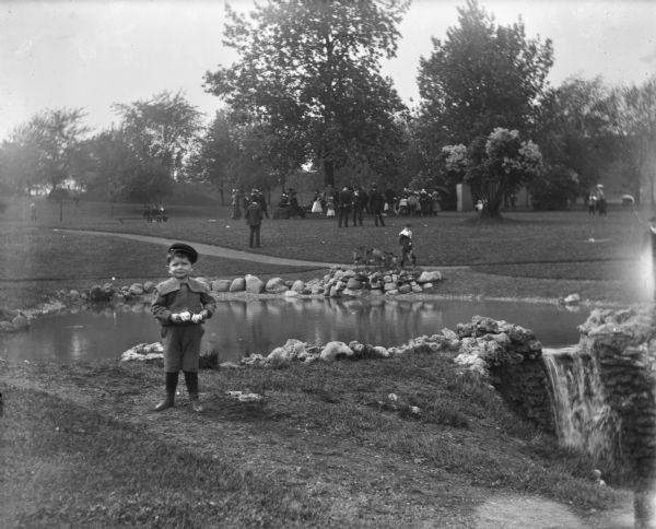 Syl standing in front of a small pond in Reservoir Park. A large group of people is gathered in the background around a tree.