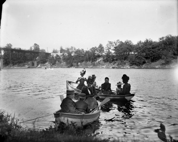 View from shoreline of Millers, Syl and others in two boats on the Milwaukee River. In the background is a boat in the middle of the river near a bridge.