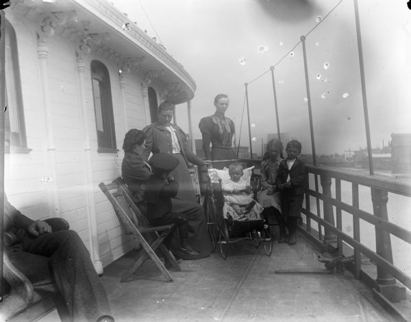 A group of children and adults sitting outdoors on the steamer "Sheboygan." Three women and four children, one in a stroller, are gathered near the railing. In the background is a body of water and on the shoreline are industrial buildings.