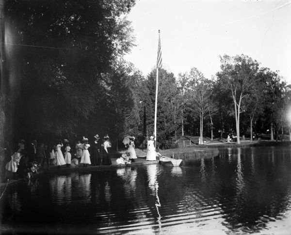 View across pond of Syl at Soldiers Home with aunt Helen standing on a landing near a boat and a flagpole. There is a large group of people gathered around the edge of the water on the left.