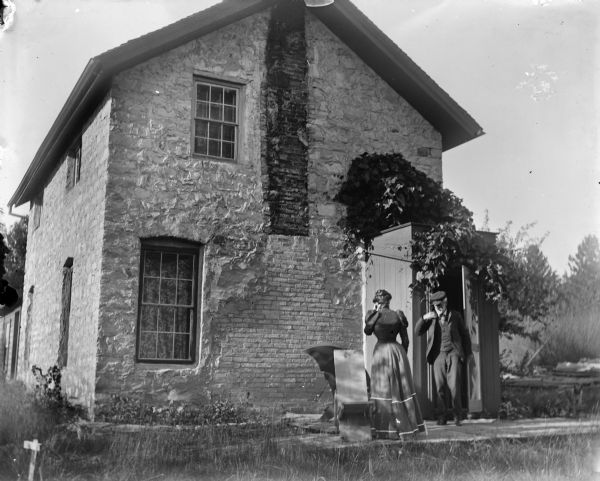 A couple in front of Grandall's house. The man and woman are standing near a shed-like entrance to the house. There are traces of an old chimney on the brickwork.