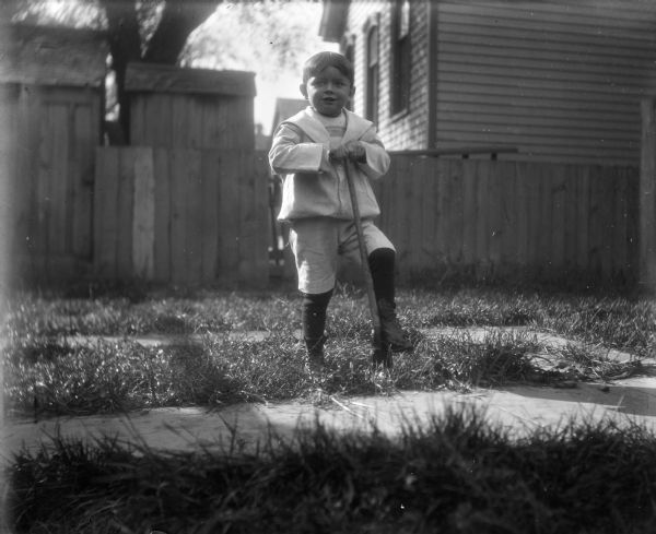 Syl leaning on a spade outside in a yard. There is a fence and a house in the background.