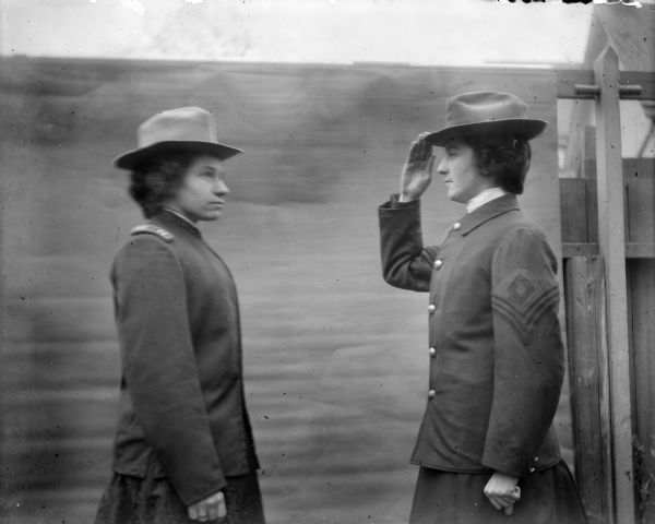 Aunt Helen and Mrs. Quill stand in front of a backdrop near a fence. They are both wearing military style uniforms including hats. The woman on the right and one is saluting the other woman on the left.