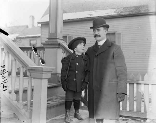 Mr. Archer stands on porch steps with his arm over Syl's shoulder. Mr. Archer has a moustache and is wearing a bowler hat and a winter coat. Snow is visible on house rooftops behind them.