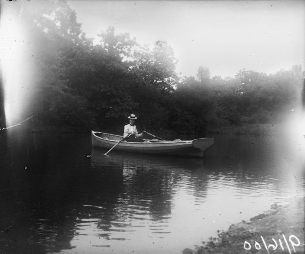 View from shoreline of a woman wearing a wide brimmed straw hat sitting in a row boat in a small body of water with the oars in her hands.