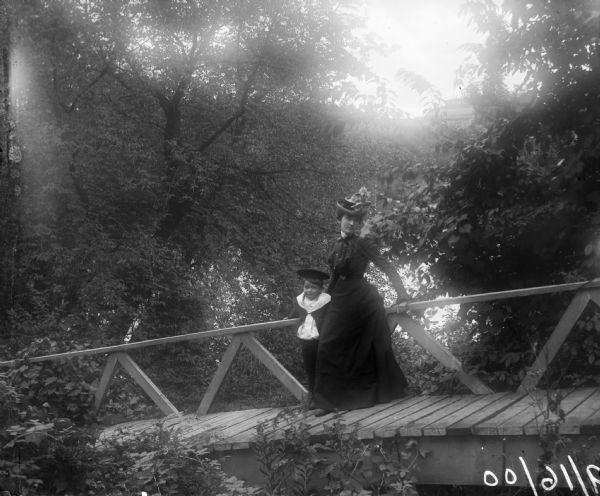 Aunt Helen and Syl stand on Gulle bridge near Gordan Place. Dense foliage surrounds them, and in the background is a large building.