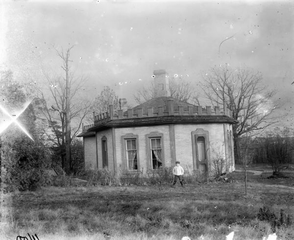 Syl standing in front of a octagonal-shaped Gordon Cottage. The edge of the roof has crenelations along it, and a brick chimney is in the middle the roof. There is overgrown grass in the foreground and trees in the background.