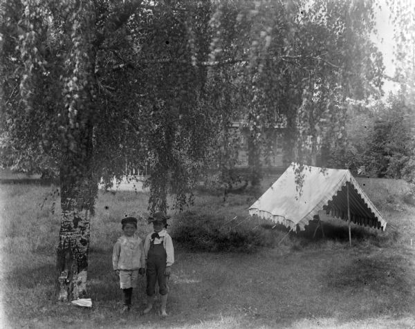 Syl and Lucia posing near a tent or open-sided awning and a willow tree. Syl is wearing pants while Lucia is wearing overalls. Both are wearing hats. In the background is a house.