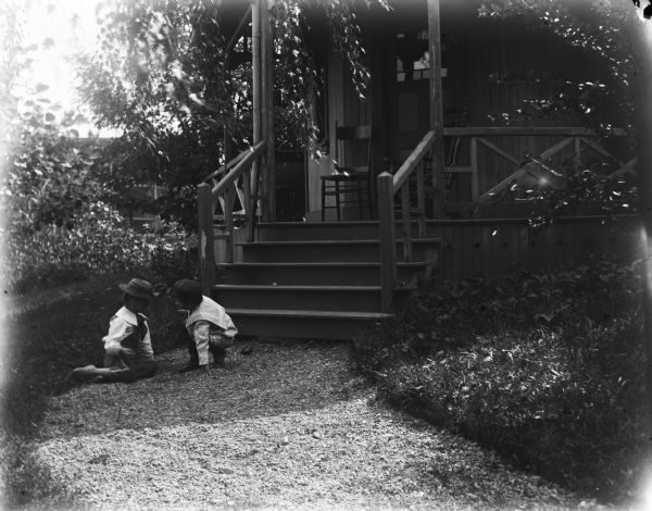 View up gravel walk of Syl and Lucia, both wearing hats, playing at the foot of steps leading up to a small porch and door of a house.