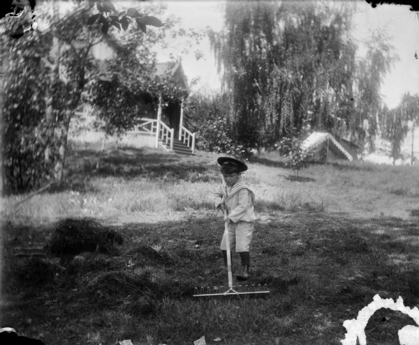 Syl raking a lawn wearing a sailor suit with a cap. In the background are steps leading up to a small porch. A small tent or open-sided awning is pitched in the yard near a tree.